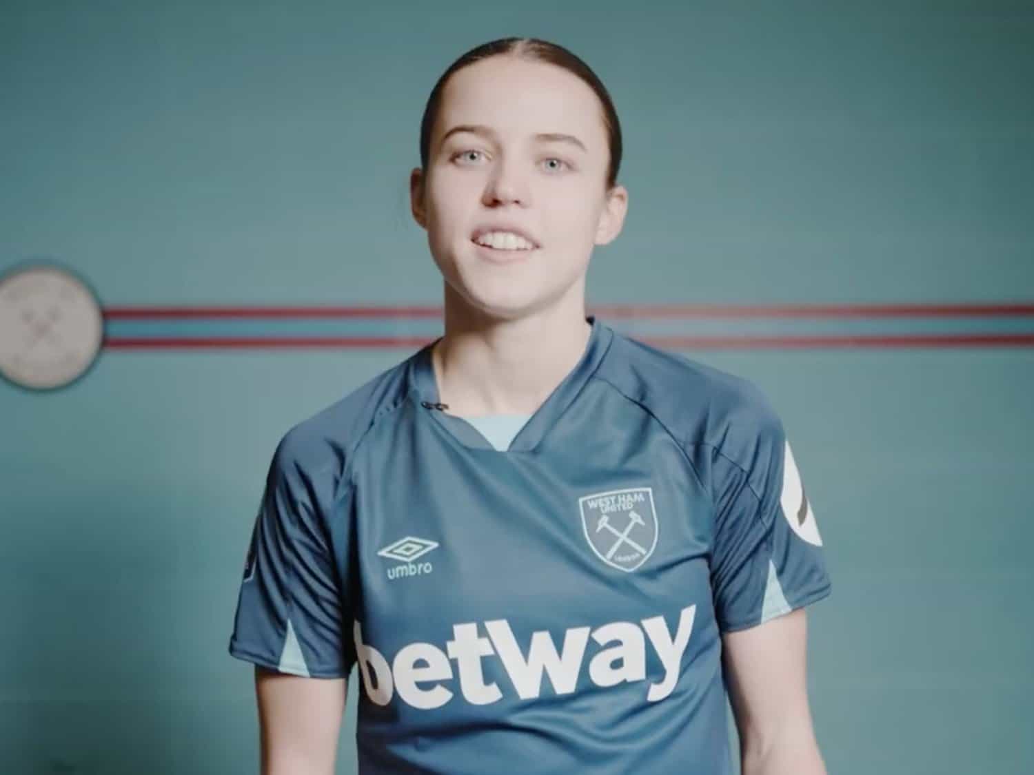 A young female athlete in a West Ham United football jersey smiling at the camera.