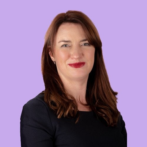 A woman with red hair and red lipstick smiling against a purple background.