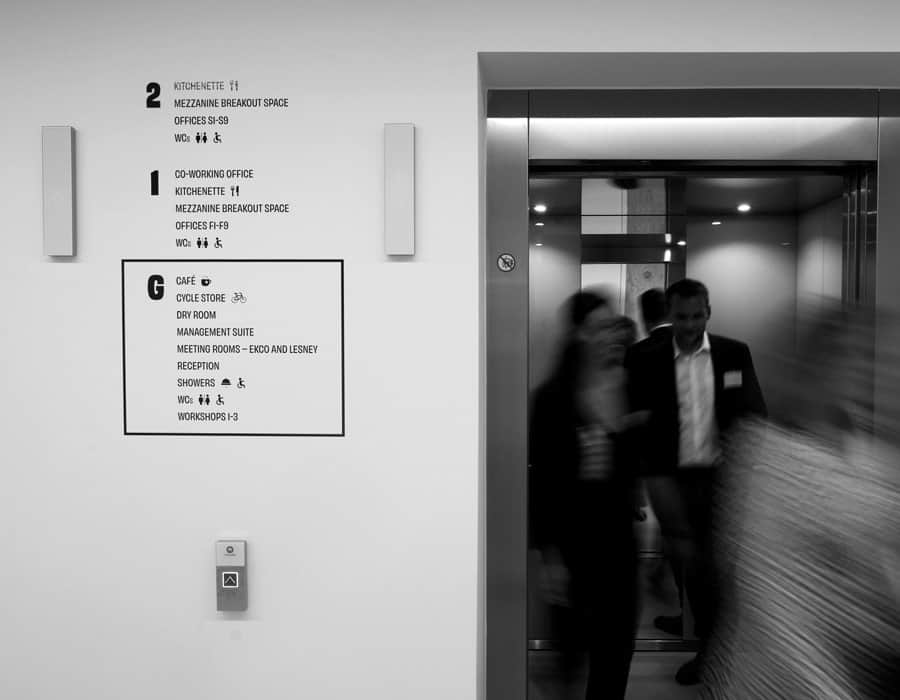 Modern office building interior with elevator and directional signage, black and white image.