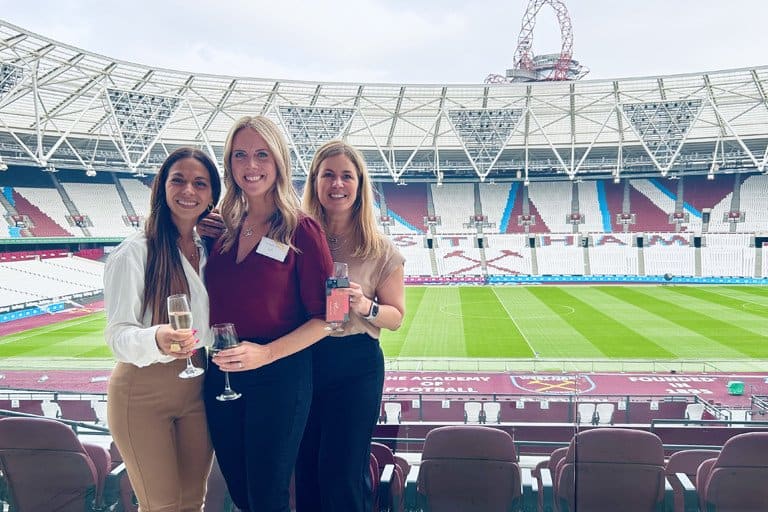 Three women at a stadium with drinks, smiling and enjoying the event.