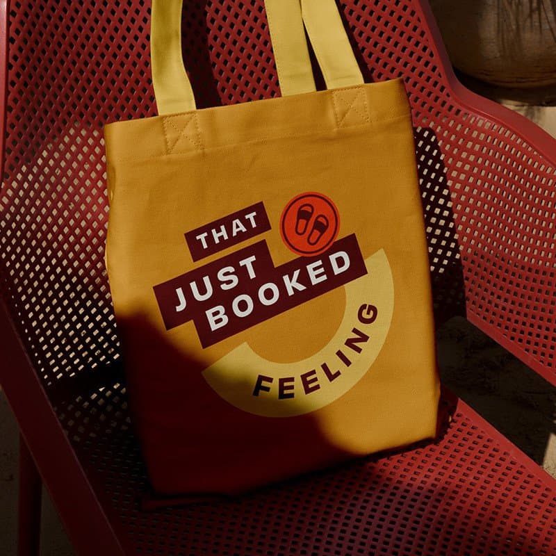 That Just Booked Feeling tote bag on red chair, promotional shopping bag, vibrant yellow color, casu.