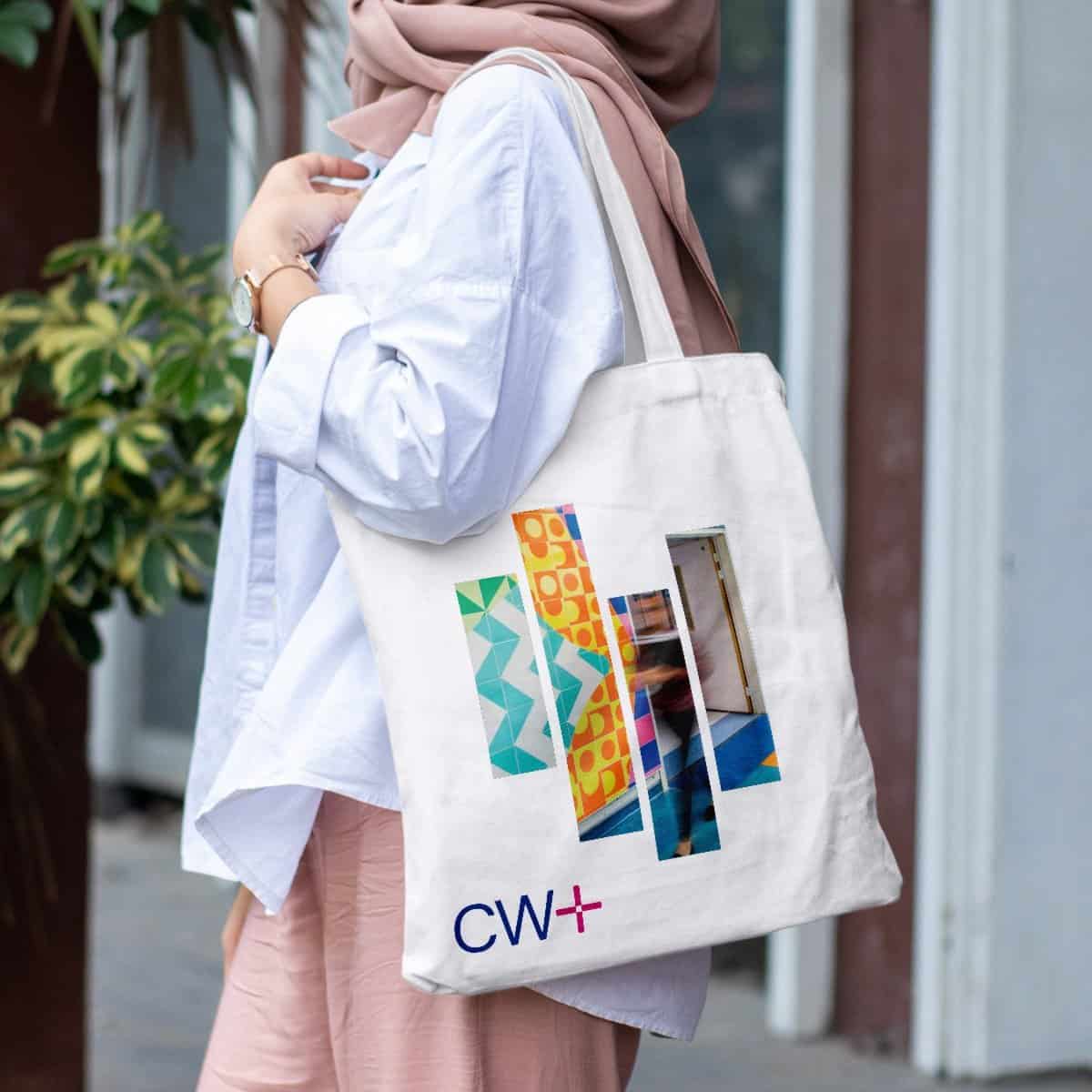 A woman holding a tote bag with colorful geometric designs and the CW+ logo, outdoor setting.