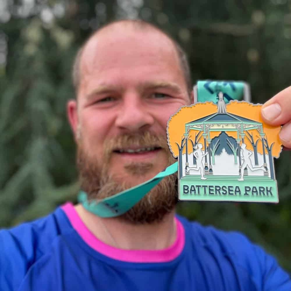 A colourful Battersea Park souvenir badge held by a smiling man outdoors in Battersea Park, London.
