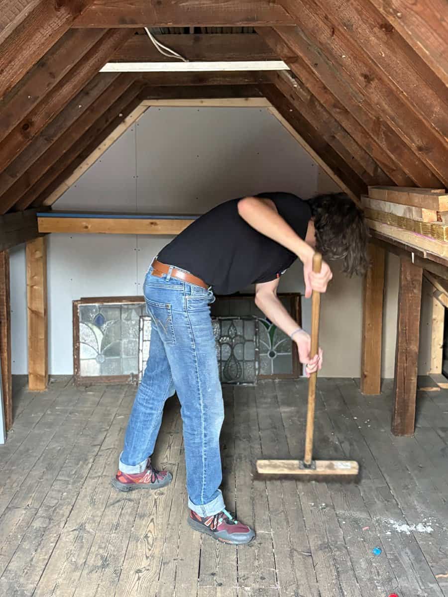 Person cleaning attic space with broom under wooden roof structure.