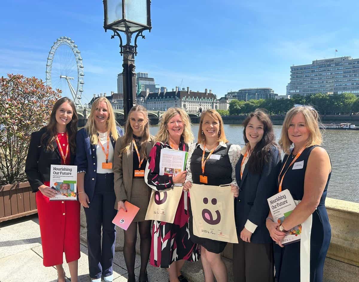 A woman and six young women standing outdoors by the river in London, holding flyers and tote bags,.
